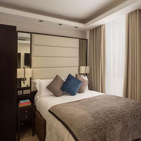 伦敦 巴比肯客房酒店 外观 照片 The image shows a cozy bedroom. It features a comfortable double bed with a light-colored duvet and decorative pillows in shades of blue and gray. The headboard is upholstered in a soft material, and there are bedside lamps on either side of the bed.