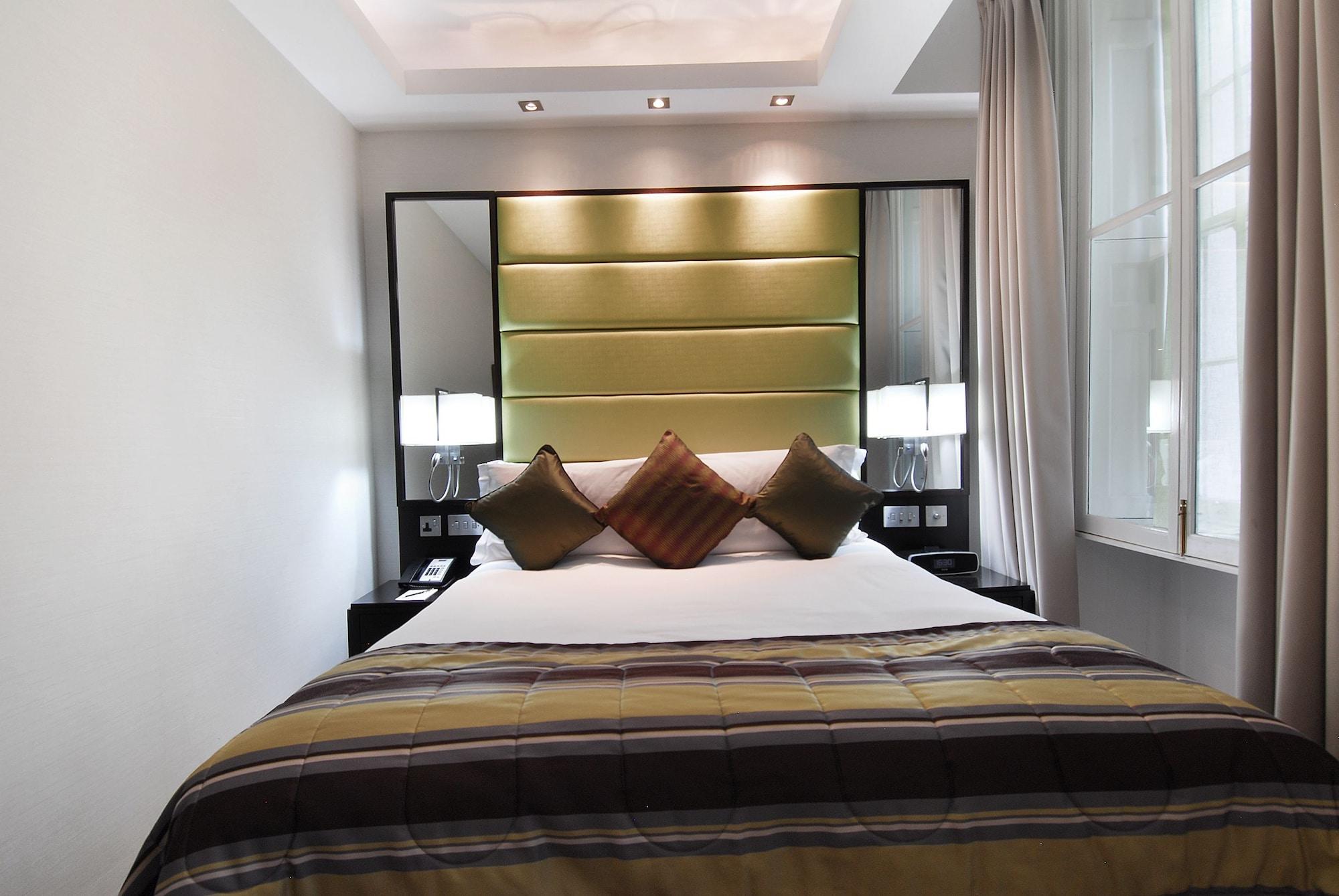 伦敦 巴比肯客房酒店 外观 照片 The photo shows a stylishly decorated bedroom. The bed is centered in the room, featuring a white comforter with a plaid pattern in shades of brown and green. There are three decorative pillows on the bed, two in shades of brown and one in dark green
