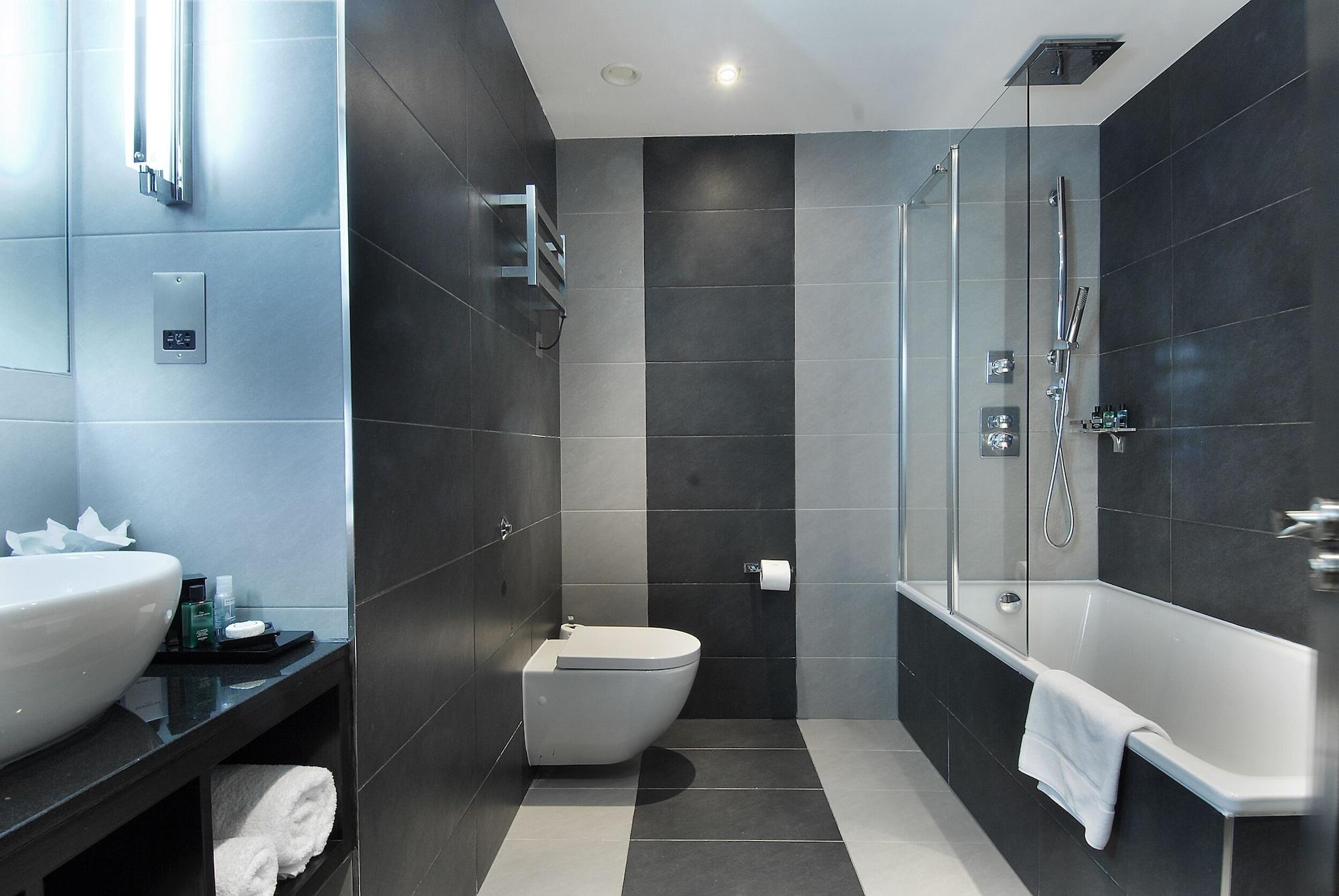 伦敦 巴比肯客房酒店 外观 照片 The photo shows a modern bathroom featuring a sleek design. The walls are tiled in a combination of dark gray and light gray, creating a contemporary look. On the left, there is a countertop with a basin and some toiletries placed on it. The middle s