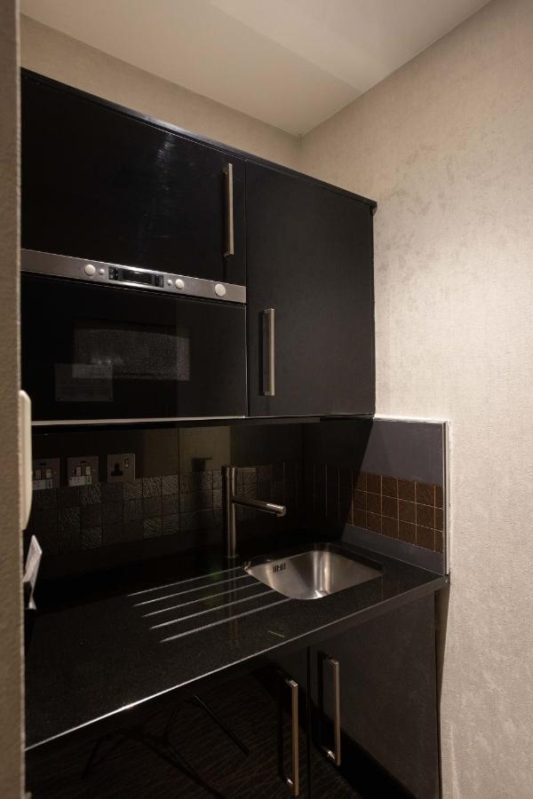 伦敦 巴比肯客房酒店 外观 照片 The photo shows a modern kitchen setup with a dark color scheme. It features:

- A built-in microwave oven situated above a countertop.
- A sleek black cabinet beneath the microwave, which likely stores kitchen items.
- A stainless steel sink placed 