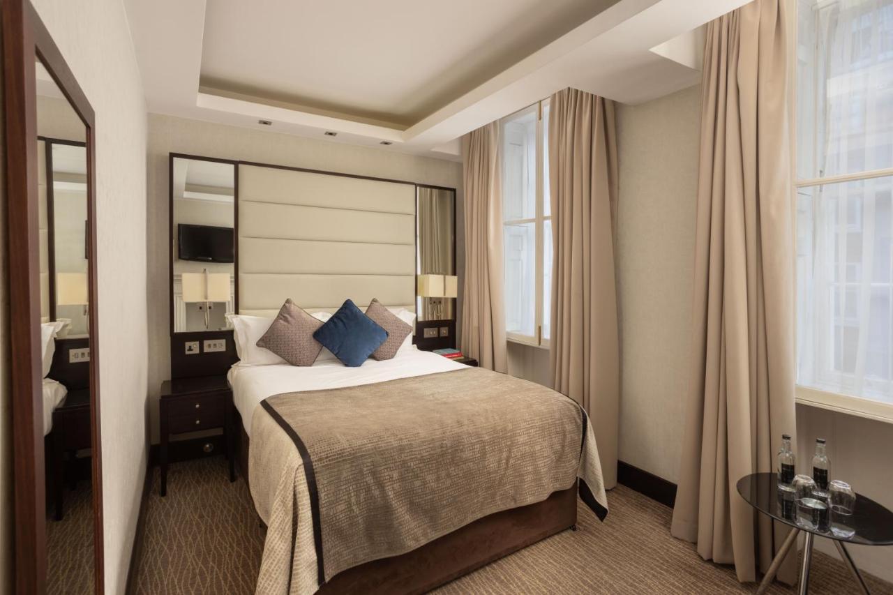 伦敦 巴比肯客房酒店 外观 照片 The photo shows a cozy hotel bedroom. The room features a neatly made double bed with a beige bedspread and decorative pillows, including a blue one. There are bedside tables on either side of the bed with lamps. The walls are painted in a soft color