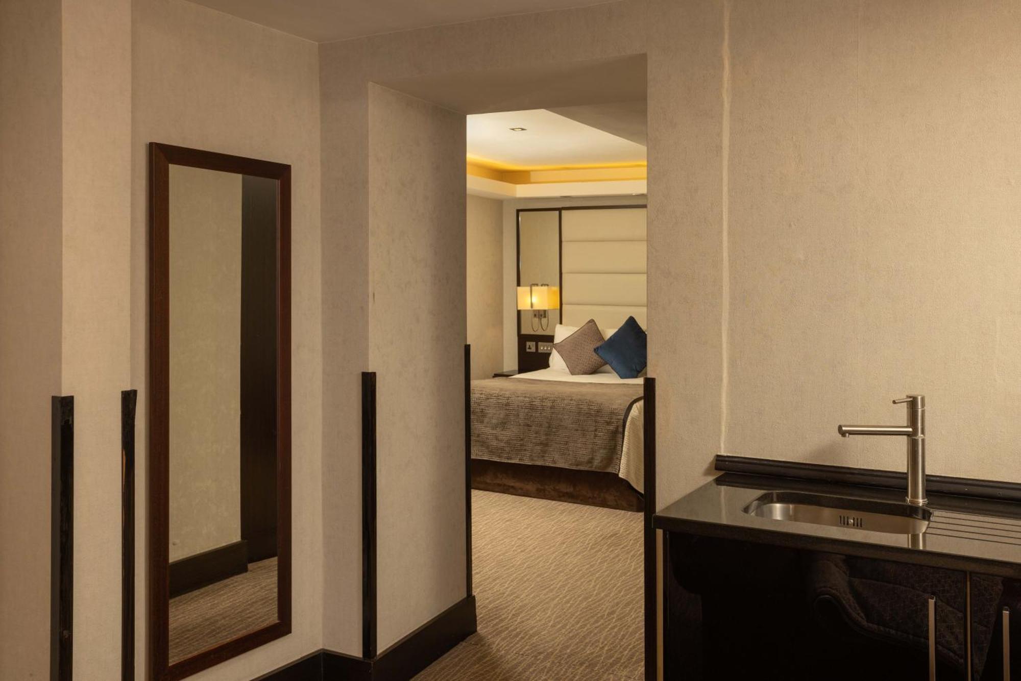 伦敦 巴比肯客房酒店 外观 照片 The photo shows an interior space that appears to be part of a hotel room or suite. There is a doorway leading into a bedroom area, where a bed with decorative pillows is visible. In the foreground, there's a tall mirror on the left side and a small 
