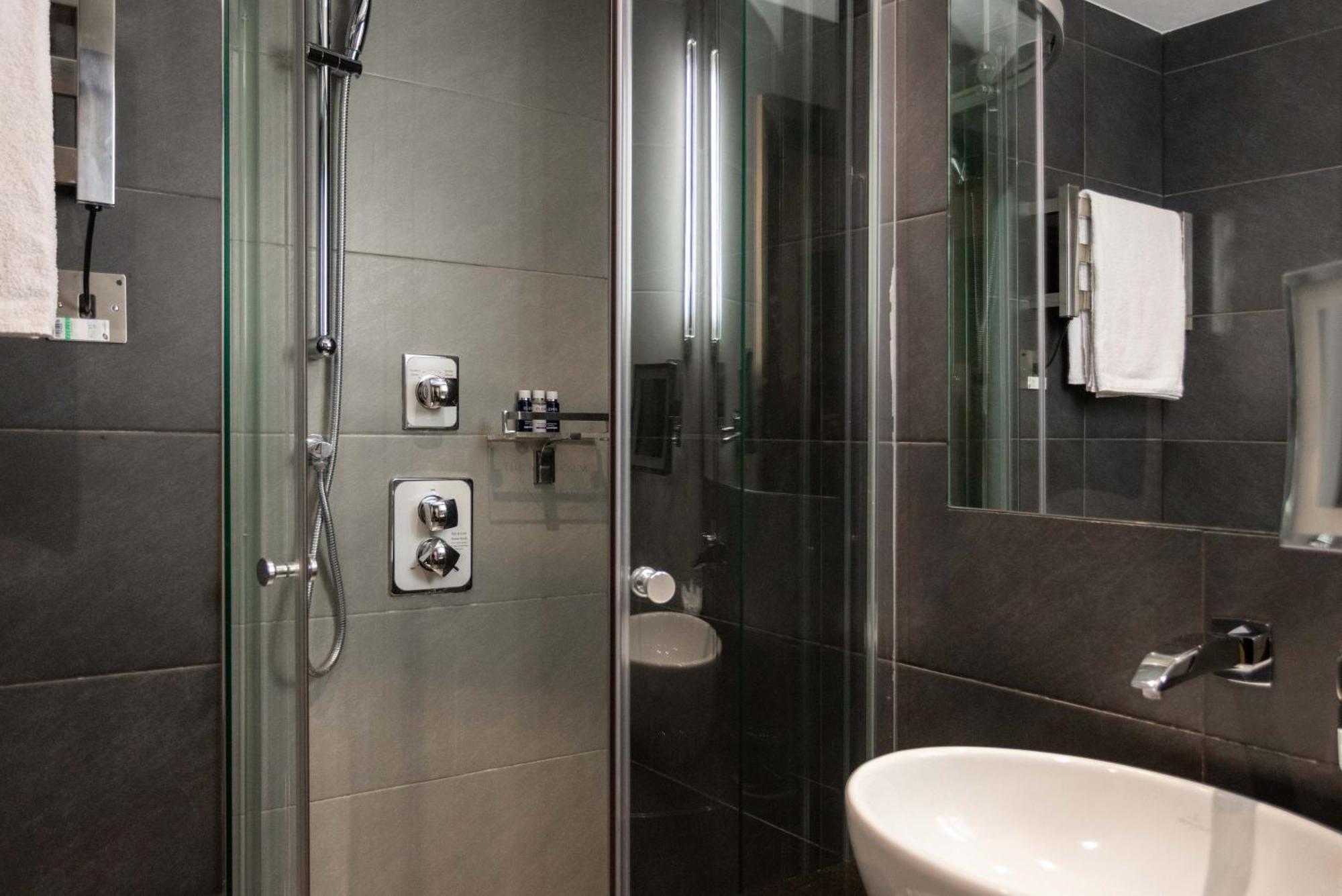 伦敦 巴比肯客房酒店 外观 照片 The photo shows a modern bathroom with a glass shower enclosure. The shower features a handheld showerhead and various controls mounted on the wall. The walls are tiled in dark, sleek tones, contributing to a contemporary aesthetic. There is a round 