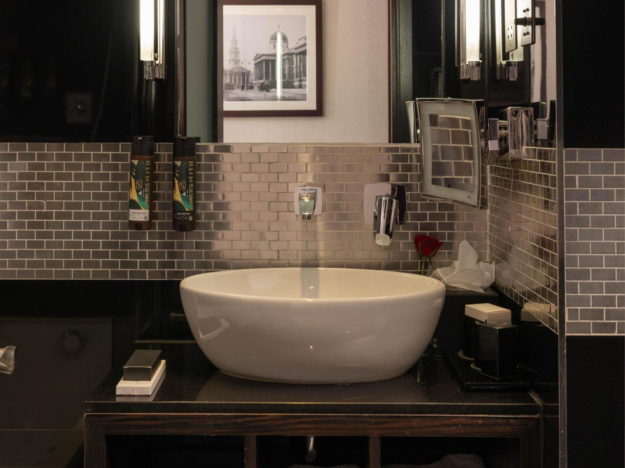 伦敦 巴比肯客房酒店 外观 照片 The photo shows a modern bathroom with a stylish design. It features a large, round white sink placed on a dark wooden vanity. The walls are adorned with a mix of gray and silver tile, creating a sleek and contemporary look. Above the sink, there is 