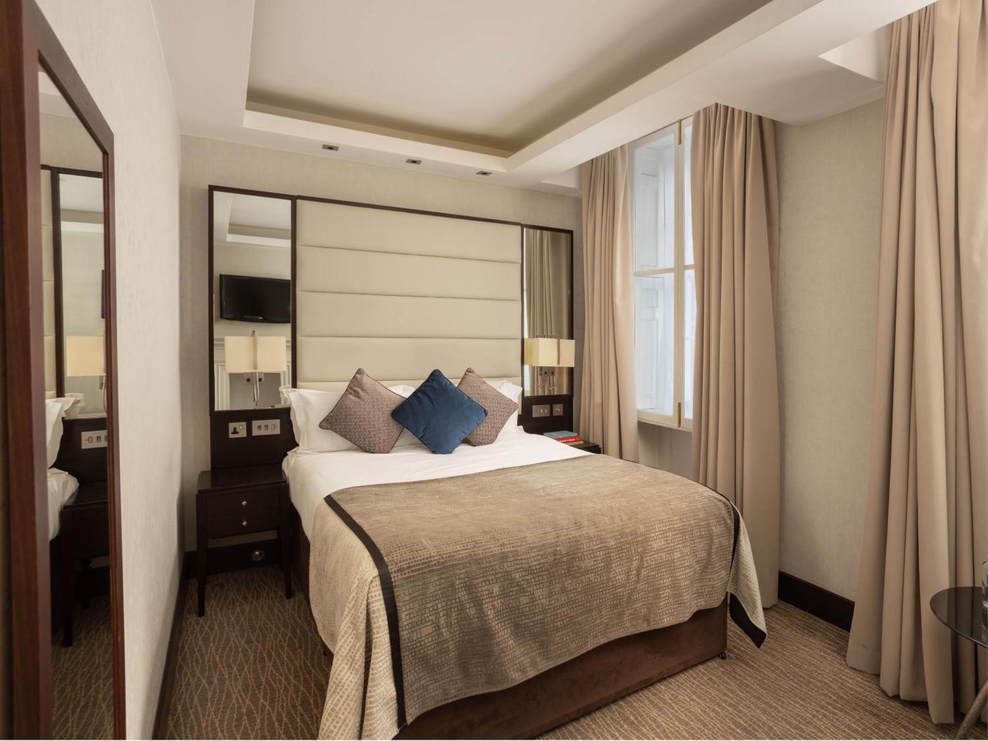 伦敦 巴比肯客房酒店 外观 照片 The photo shows a cozy hotel room with a bed as the focal point. The bed is made with neutral-colored linens and has several pillows, including a decorative blue one. There are bedside tables on either side of the bed, each with a lamp. A large mirro