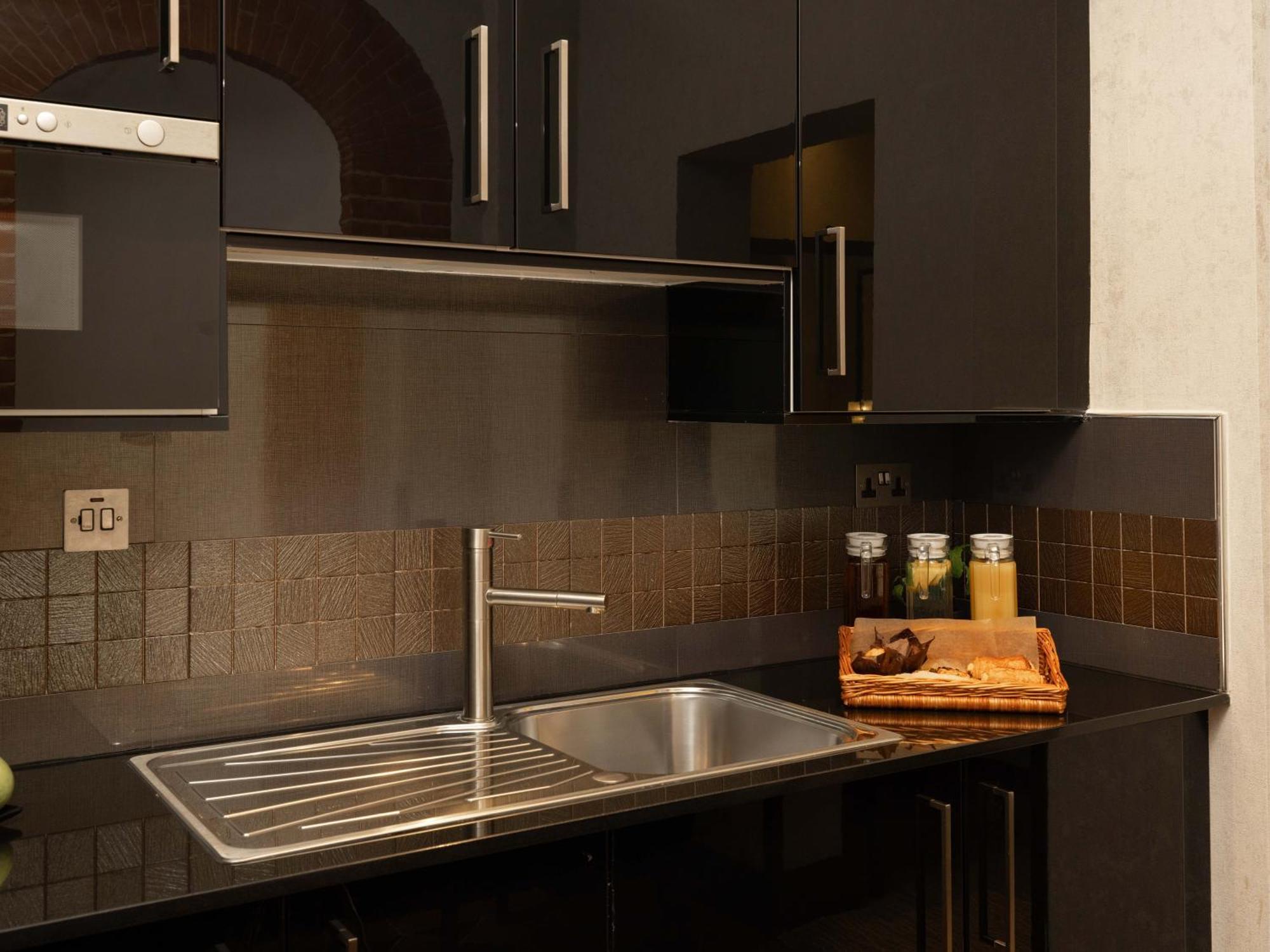 伦敦 巴比肯客房酒店 外观 照片 The photo shows a modern kitchen with a sleek design. It features dark cabinetry with a glossy finish and stainless steel handles. There is a stainless steel sink with a simple faucet, and a countertop made of black material. Above the sink, there ar