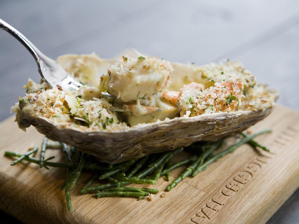 伦敦 巴比肯客房酒店 外观 照片 The photo shows a large oyster shell filled with a mixture that appears to include seafood, herbs, and breadcrumbs. The dish is presented on a wooden board with some greenery, possibly a type of seaweed or garnishing. A fork is placed beside the oyst