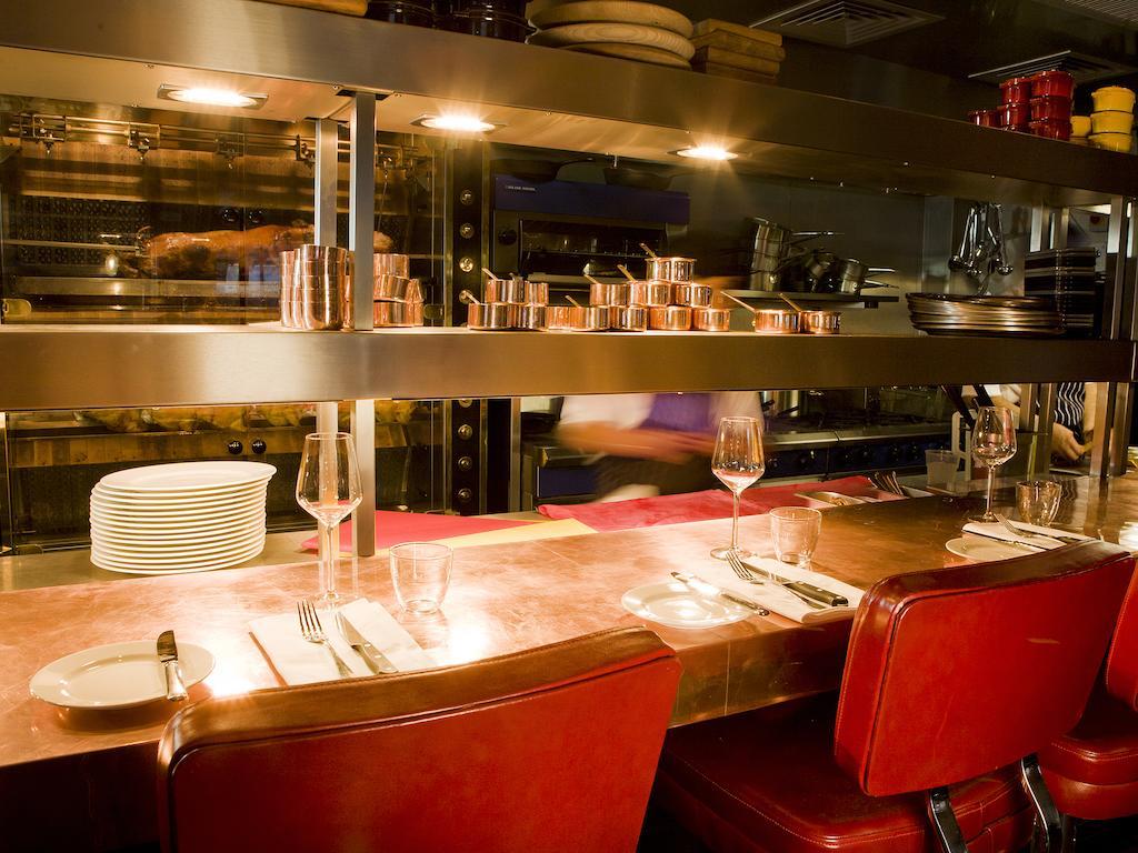 伦敦 巴比肯客房酒店 外观 照片 The photo shows a restaurant interior featuring a bar or counter area. There are red chairs arranged around a copper-topped table where dining utensils and plates are set. Above the counter, there are shelves displaying various cooking pots and kitch
