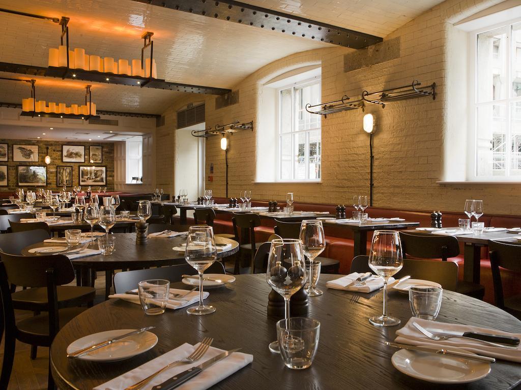 伦敦 巴比肯客房酒店 外观 照片 The photo shows a restaurant interior featuring a warm and inviting atmosphere. It has several round wooden tables set for dining, each with neatly arranged plates, utensils, and glasses. The lighting is soft, with overhead fixtures providing a cozy 