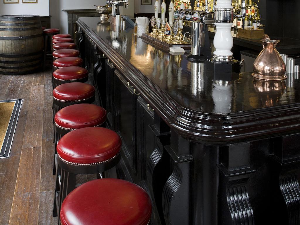 伦敦 巴比肯客房酒店 外观 照片 The photo shows a bar setting with a long, polished dark wooden bar counter. Attached to the counter are several bar stools with red leather seats. Behind the bar, there are various bottles, suggesting a well-stocked bar. The flooring appears to be w