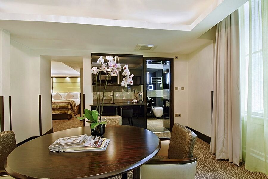 伦敦 巴比肯客房酒店 外观 照片 The photo shows a stylish hotel room or suite. In the foreground, there is a round dining table with a few magazines on it. Surrounding the table are upholstered chairs. To the right, there are large windows with light-colored curtains, allowing natu