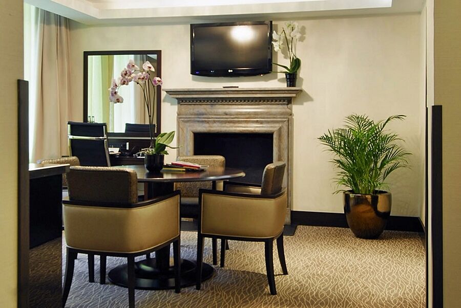 伦敦 巴比肯客房酒店 外观 照片 The photo shows a modern and stylish room with a dining or meeting area. There is a round table surrounded by four upholstered chairs. A flat-screen television is mounted on the wall above a decorative fireplace. To the side, there is a mirror reflec