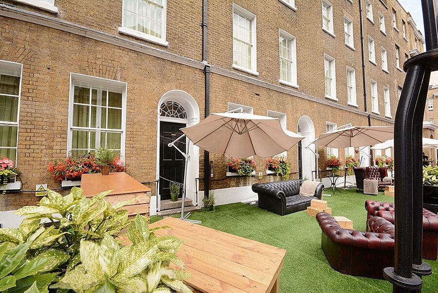 伦敦 巴比肯客房酒店 外观 照片 The photo depicts an outdoor area of a building with a brick facade. There are several large windows visible, along with a wooden door. The setting features outdoor furniture, including couches in a dark leather finish and wooden tables. Large umbrel